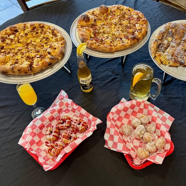 Three pizzas, two with savory toppings and one dessert pizza, with drinks and donuts on a black table.