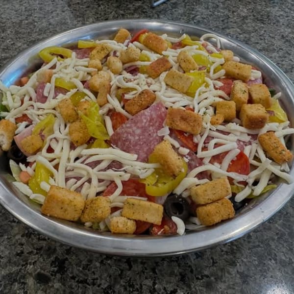 A large salad with pepperoni, salami, cheese, peppers, olives, and croutons in a metal bowl.