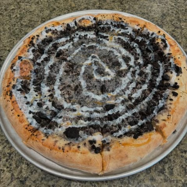 A dessert pizza with a spiral of white icing and crushed dark cookies on a metal pan.